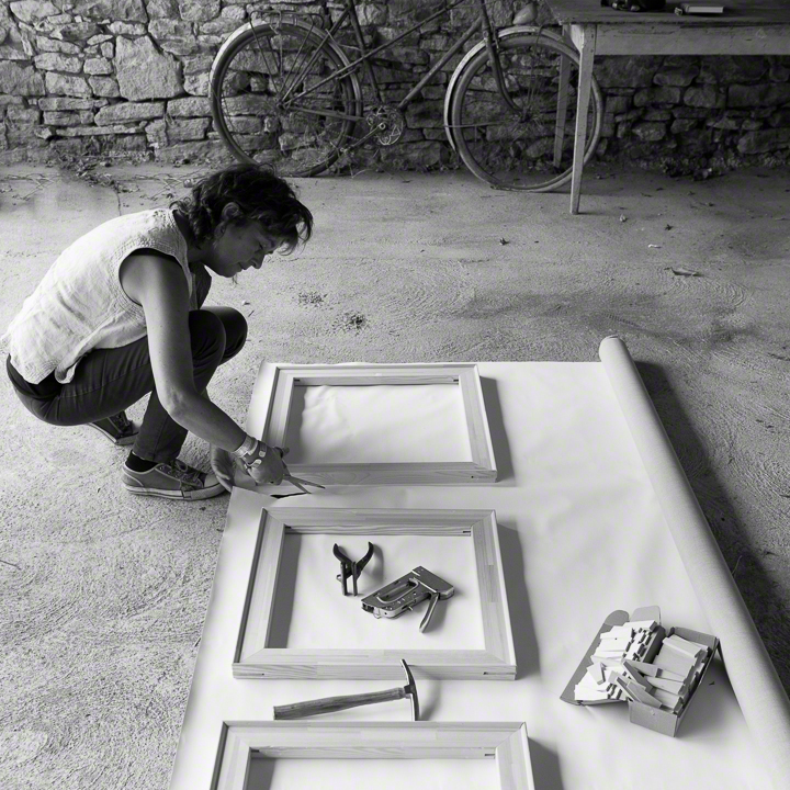 Woman, Catherine Forshall, crouching to cut canvas with scissors, on cement floor, using canvas stretcher as as template, stone wall, bicycle and table in background